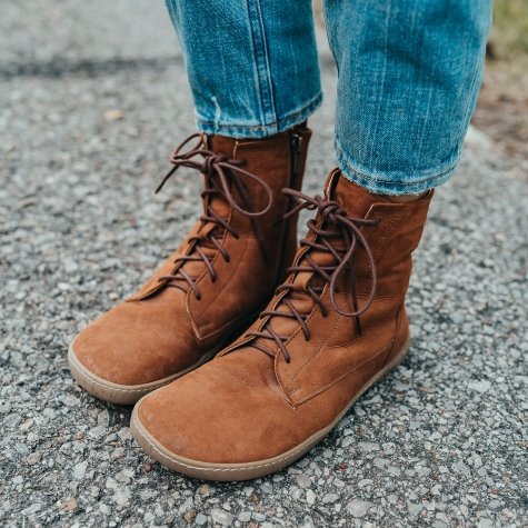 A pair of feet from the shins down wearing jeans and Shapen Cozy barefoot lace up boots with a side zipper and zero drop outsole