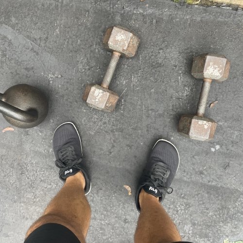 Top down view of a pair of feet wearing Freet Vibe barefoot shoes with a kettle bell and dumbbells laying on the ground around the shoes.