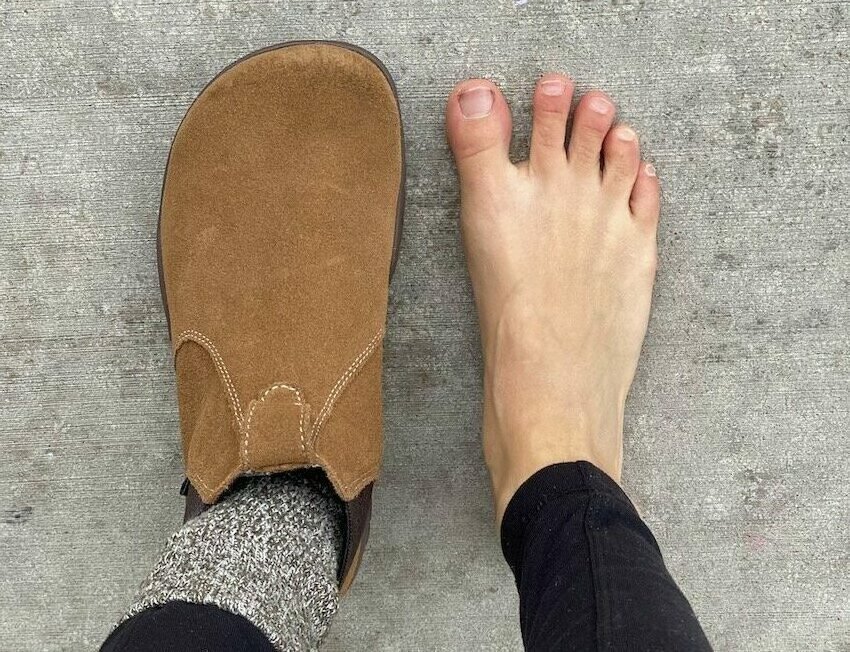 a top down view of a pair of feet standing on concrete. Left foot is wearing Lems Chelsea boots in Cedar, right foot is bare, showing that the boots have an anatomical foot shape