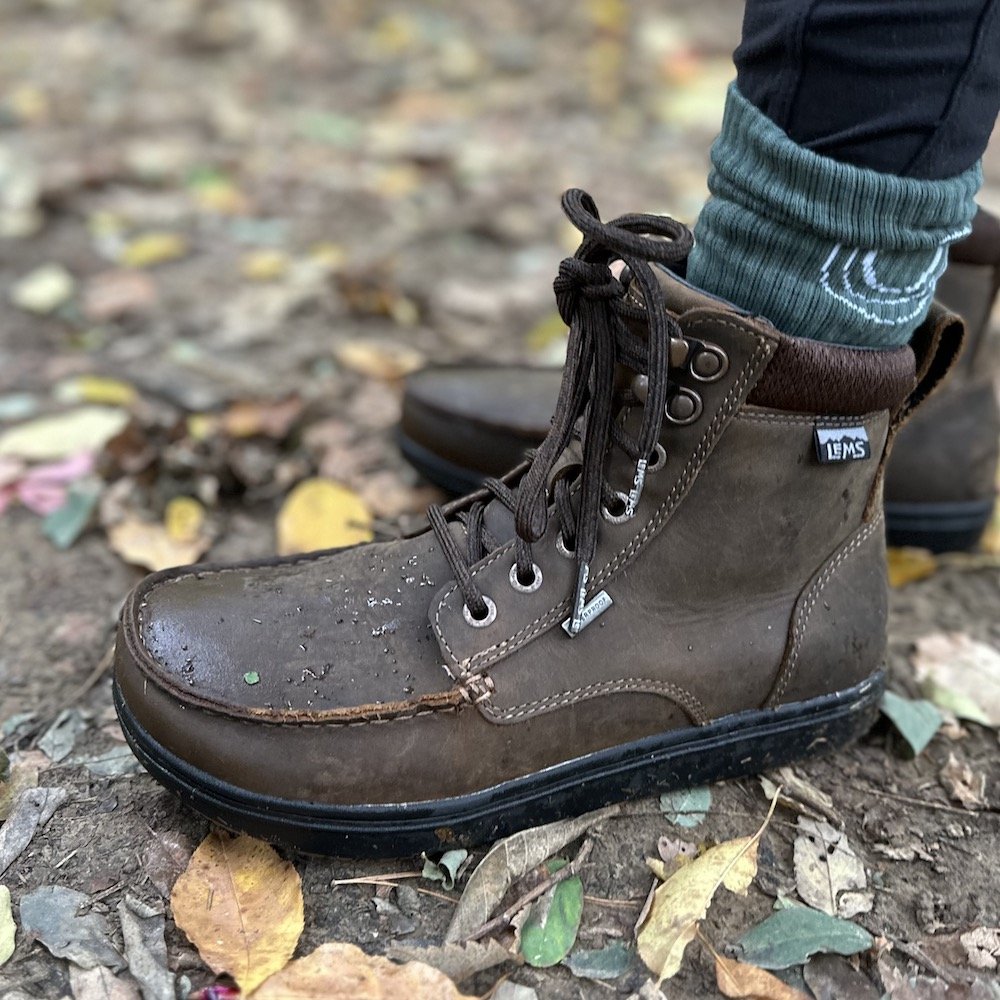 a close up side view of a pair of feet standing in a forest in the rain with leaves on the ground wearing Dakota brown Lems barefoot waterproof Boulder boot grip with a non slip sole