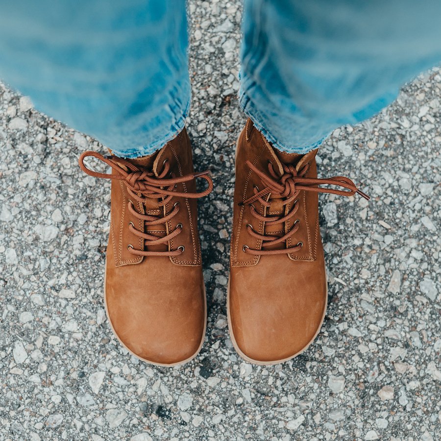 Top down ariel view of a person in jeans standing on an exposed aggregate road wearing brown Shapen Barefoot Urbaneer Boots for wide feet. The squared shape toe box allows for proper foot form.