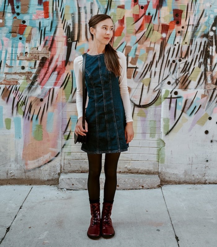 A woman standing in front of a graffitied wall, looking off to the side wearing a denim dress, tights, and red burgundy wide toe box barefoot Shapen Maverick combat boots.