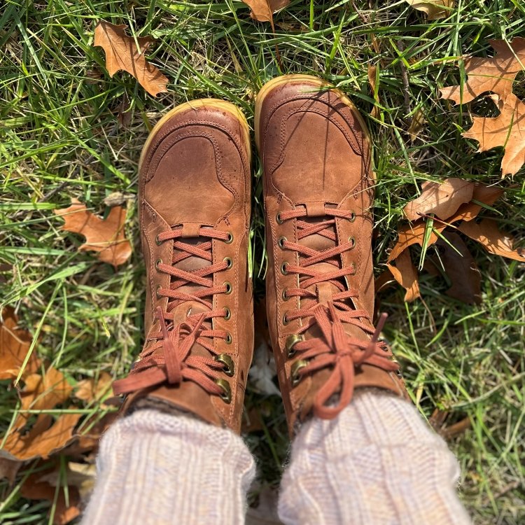 A person standing with their feet parallel in Vivobarefoot Gobi leather warmlined barefoot winter boots. There are cream colored chunky knit leg warmers peeking out over the warm brown colored leather boots