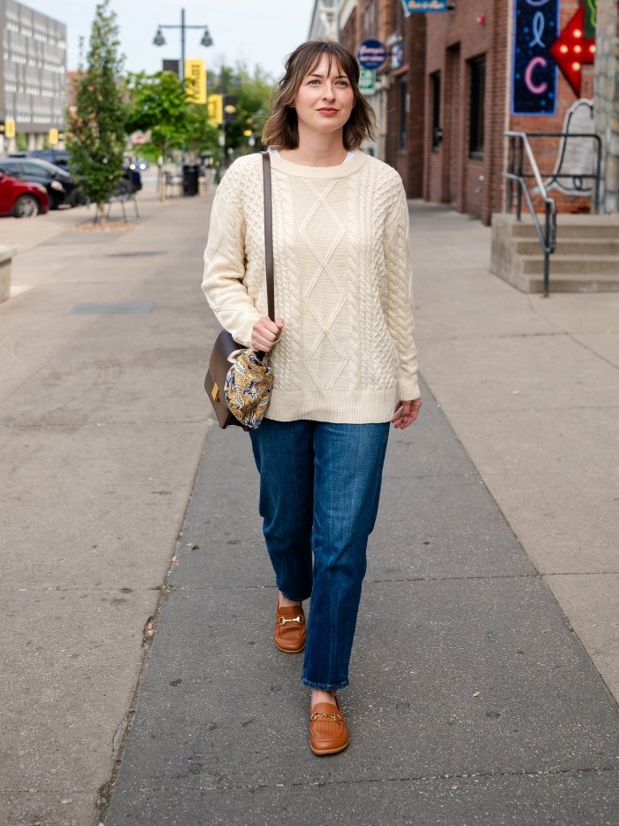 Full body view of a woman in jeans, a cable knit cream sweater, and a pair of Be Lenka Divella barefoot loafers in cognac walking on a sidewalk downtown.