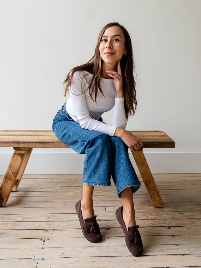 Full body image of a woman sitting on a wooden bench with her legs crossed and her elbows resting on her thighs. She has on a pair of brown suede Fiorela barefoot loafers from Be Lenka