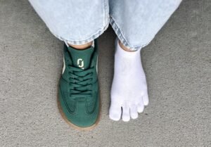 A top down view of a pair of feet. One foot is wearing toe socks and the other is wearing an Ohne Project vegan barefoot sneaker that is dark green
