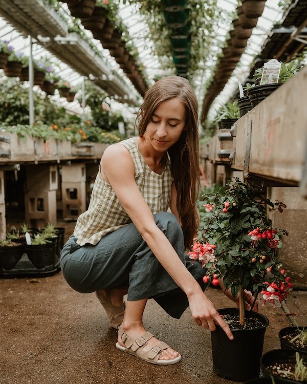 Anya crouching down in a garden center wearing linen clothes, holding a plant, with Mukishoes Nazare barefoot sandals on her feet.