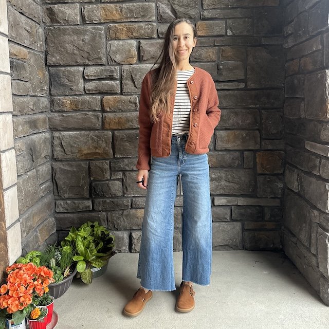 Full body view of a woman with long brown hair wearing wide leg jeans, a striped shirt, and a fuzzy brown fleece.