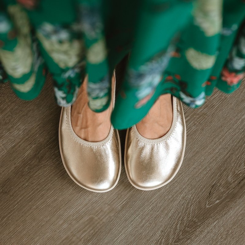 A woman in a green floral skirt with a pair of gold Groundies Lily barefoot ballet flats peeking out from under.