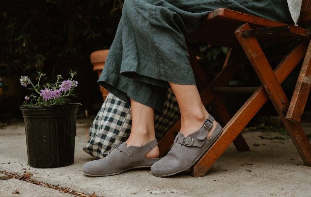 Close up knee down view of a person wearing linen pants and Mukishoes Soca clogs sitting outside on a wooden folding chair with flower pots nearby.