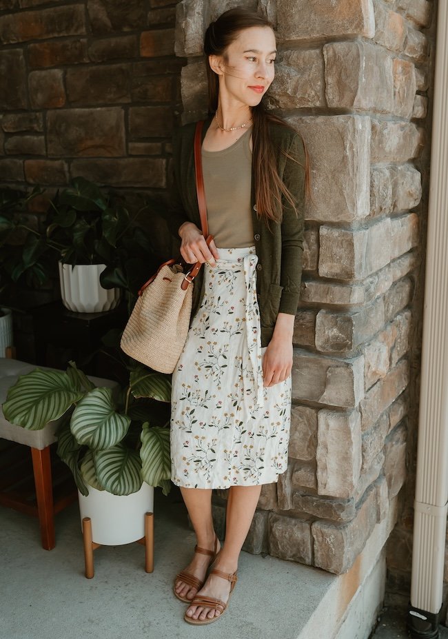A woman leaning against a rock pillar wearing a floral skirt and Be Lenka Summer barefoot brown leather sandals