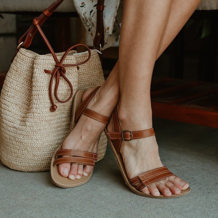 A pair of feet crossed at the ankles with a woven bag behind them. On the feet are Be Lenka Summer brown leather barefoot sandals with adjustable velcro straps