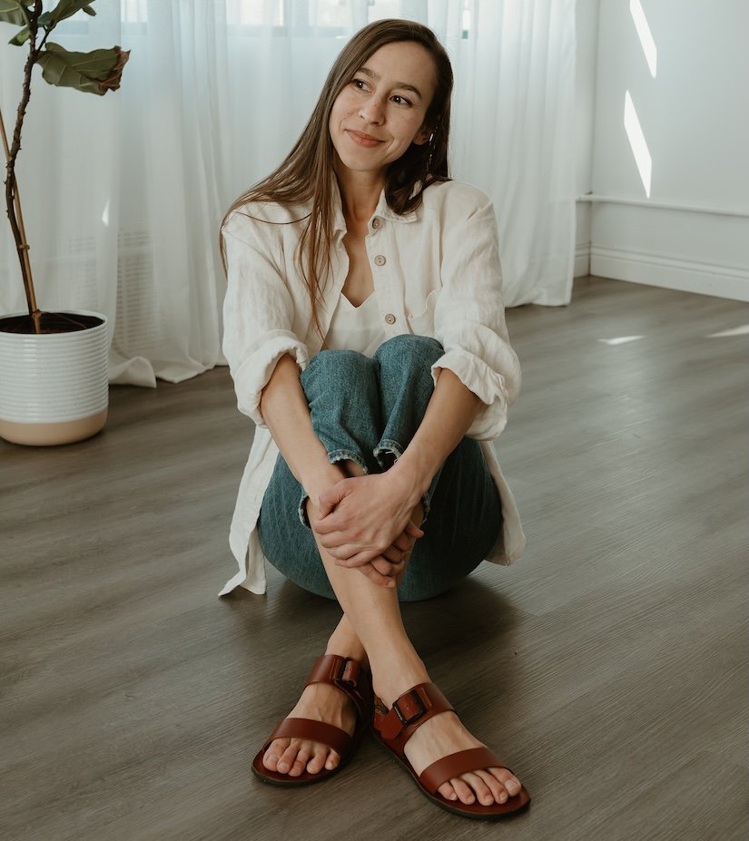 A woman sitting on the wood floor wearing denim and Groundies Kos brown leather barefoot sandals