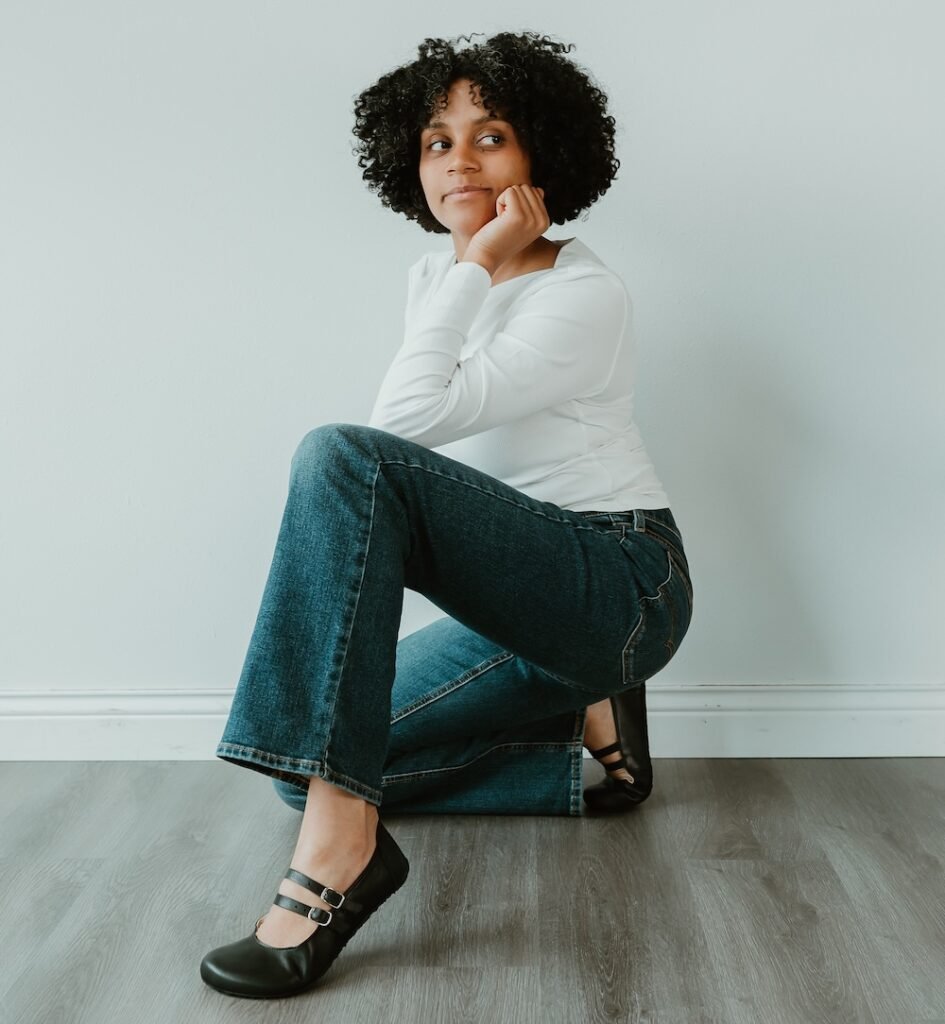 A woman with curly hair wearing a white tee shirt and jeans and black Jenon Leather barefoot mary janes, smiling at the camera