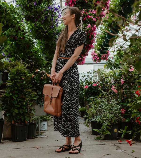 A woman standing in a greenhouse wearing a black floral dress and black leather Laboo barefoot sandals with her face turned away