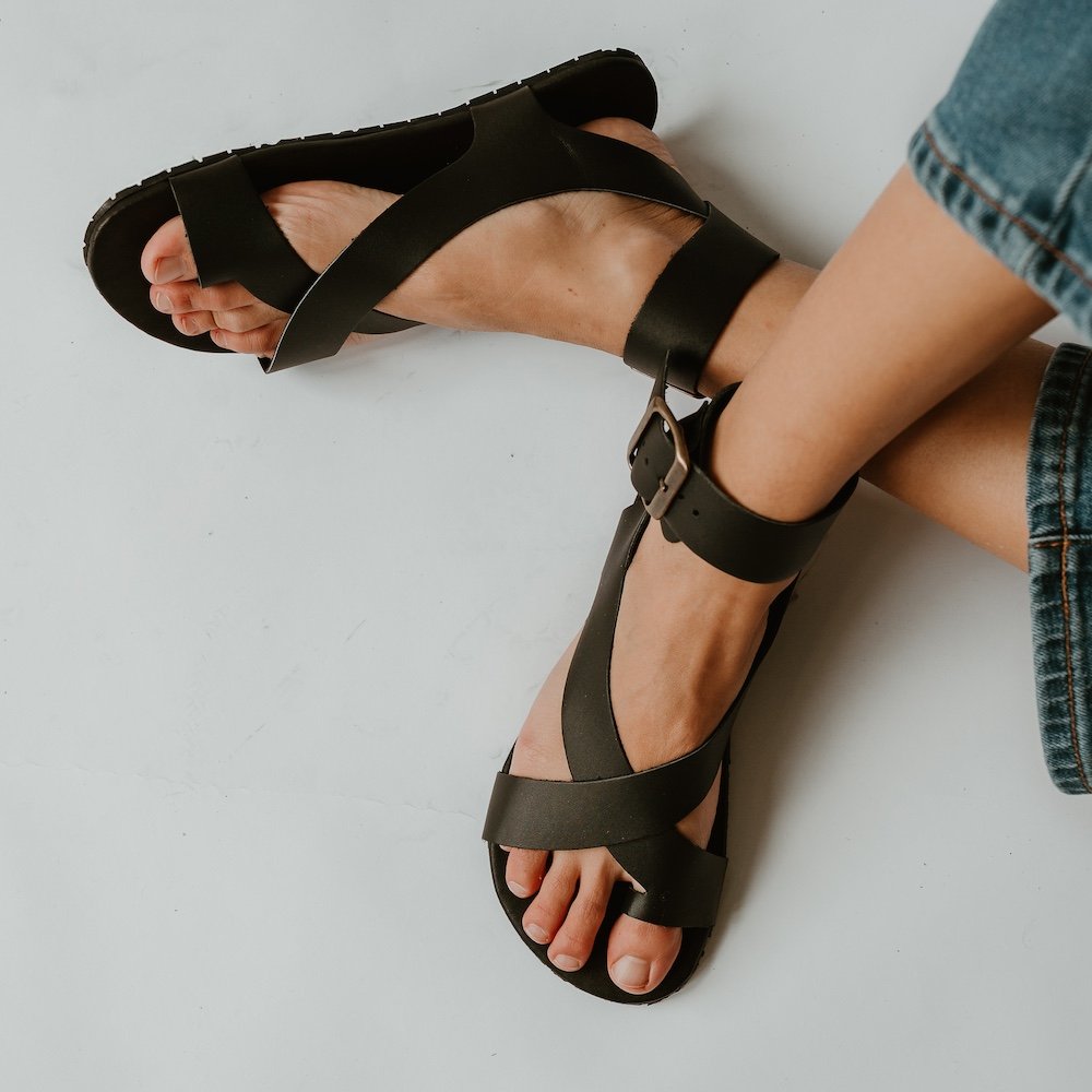 A top down view of a pair of feet over a white background wearing black leather Mukishoes Ibiza thick-strapped barefoot sandals