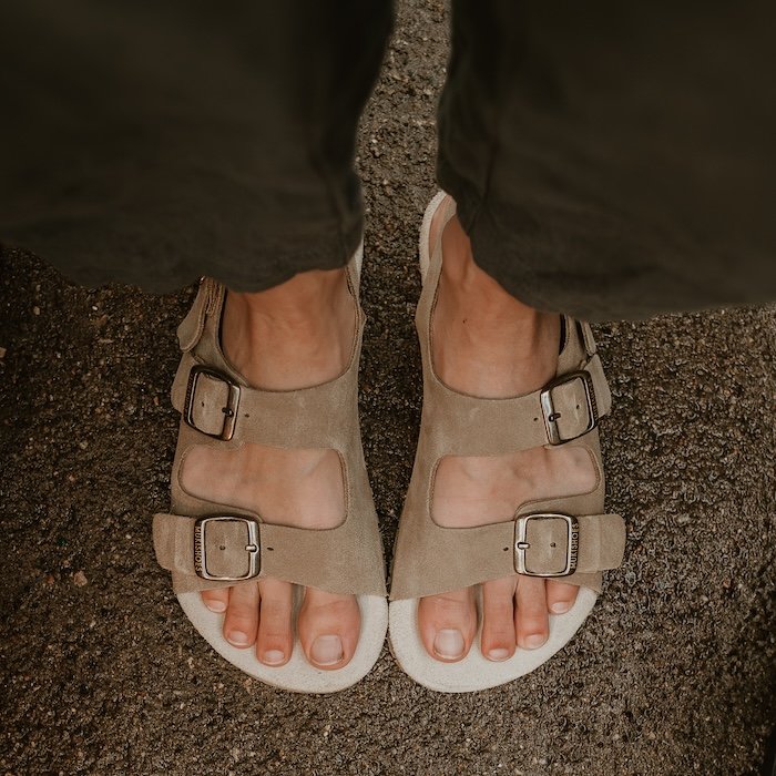 A top down view of a pair of feet standing on concrete wearing light brown suede buckle strap barefoot sandals called the Mukishoes Nazare