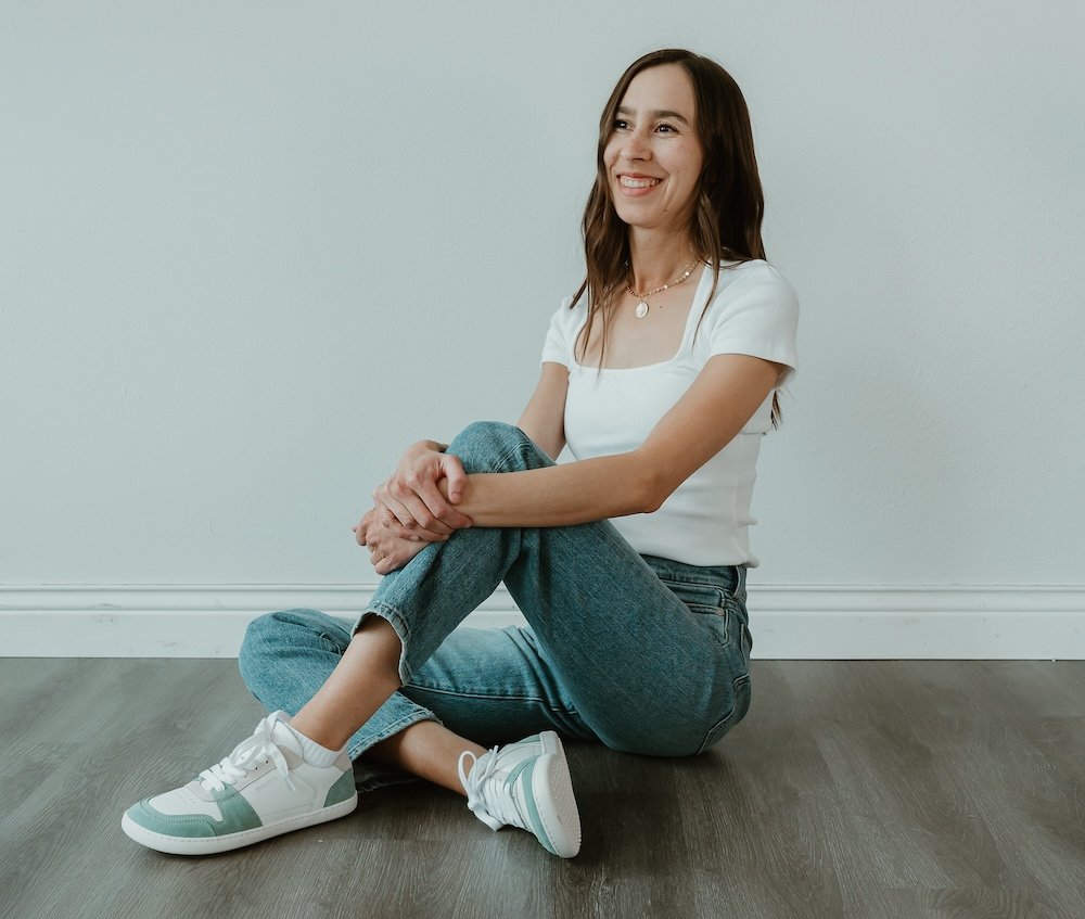A woman sitting on the wood floor wearing jeans, a white shirt, and Shapen ReWind Jade barefoot sneakers
