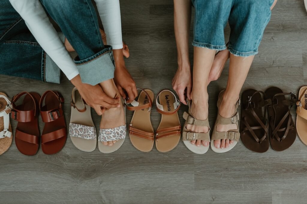 A row of stylish cute barefoot sandals for women in different shades of brown and tan from brands like Mukishoes, Be Lenka, Laboo, Zeazoo and Groundies with two pairs of legs and feet each putting a pair on