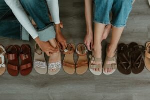 A row of stylish cute barefoot sandals for women in different shades of brown and tan from brands like Mukishoes, Be Lenka, Laboo, Zeazoo and Groundies with two pairs of legs and feet each putting a pair on