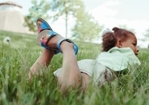 A child laying on her stomach in grass and dandelions with her feet up behind her crossed. She is looking off to the side and on her feet are Zeazoo Ariel kids barefoot summer sandals made of a bright blue leather with multicolored hearts on the strap.