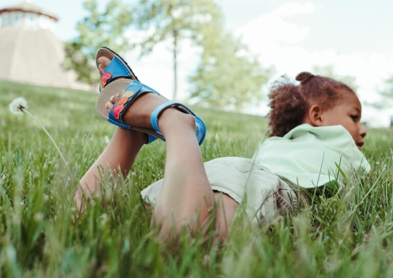 A child laying on her stomach in grass and dandelions with her feet up behind her crossed. She is looking off to the side and on her feet are Zeazoo Ariel kids barefoot summer sandals made of a bright blue leather with multicolored hearts on the strap.