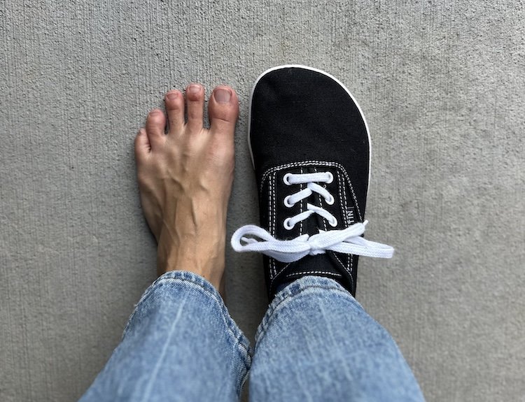 Top down view of a woman with 1 bare foot and 1 foot wearing Whitin canvas barefoot sneakers like Keds, but with a wide anatomical toe box