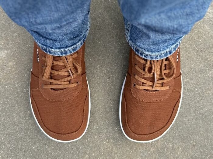 Top down close up of a man's feet wearing jeans and Whitin canvas barefoot sneakers, with matching laces and a wide toe box
