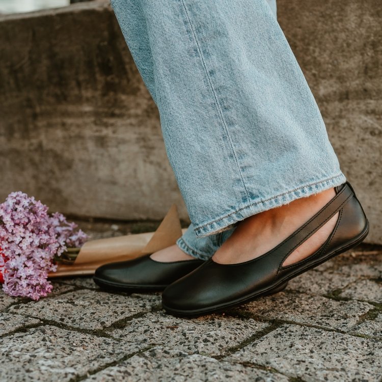 Side close up of a person's feet in Be Lenka Belissima balck cut out barefoot flats being worn with light colored wide leg jeans. There is a bundle of purple flowers peeking out in the background and the mossy stones are underfoot. The barefoot flats zero drop outsole is in the forefront of the image.