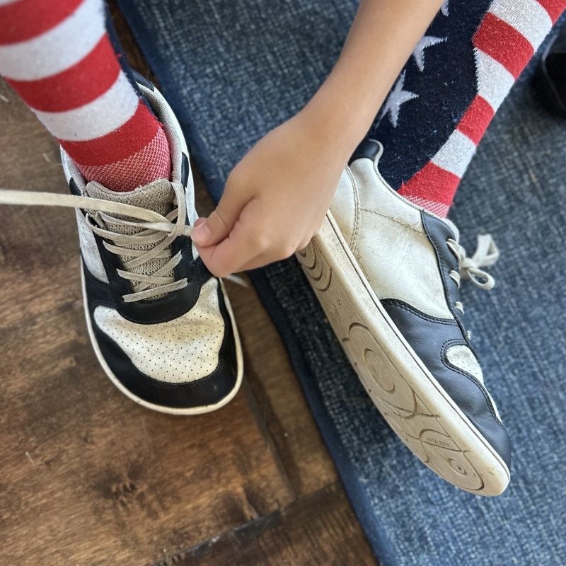 A child tying the laces on a pair of black and white leather wide toe box sneakers from Be Lenka. One foot is laying open to the side showing the zero drop outsole, and the other is flat on the floor highlighting the wide anatomical toe box.