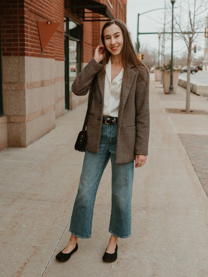 Full body view of a woman in cropped mid tone jeans, a white silky dress shirt, and a menswear inspired plaid blazer standing on the sidewalk tucking the hair behind her ear. On her feet are classy black Be Lenka Sophie barefoot ballet flats with a wide toe box.