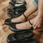A line of black barefoot ballet flats with a persons feet being slipped into one pair. They all have a wide toe box and flexible flat outsole