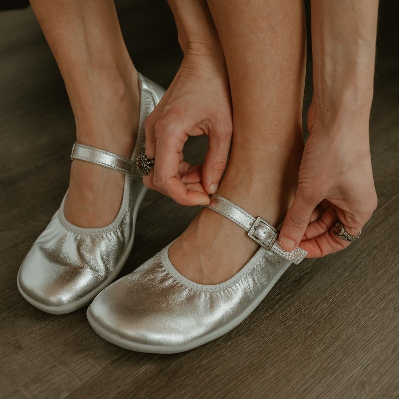 Close up of a person buckling the strap on a pair of pretty silver Groundies Amber mary jane flats.
