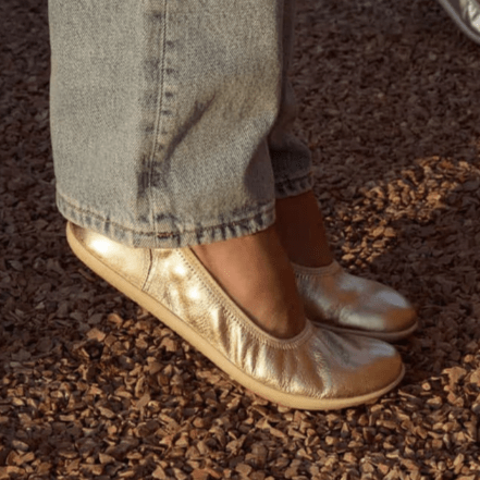 Groundies Lily 3.0 barefoot ballet flats being worn by someone standing on their tip toes. The champagne metallic leather is shining in the evening light, worn with a pair of light colored jeans