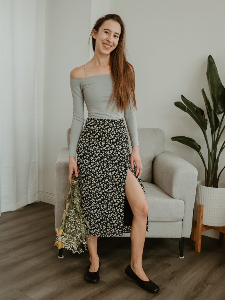 Full body photo of a woman in an off-the-shoulder gray top and a floral skirt with a slit. She is holding a bouquet of flowers and smiling at the camera. On her feet are Groundies Lily barefoot ballet flats in black leather.