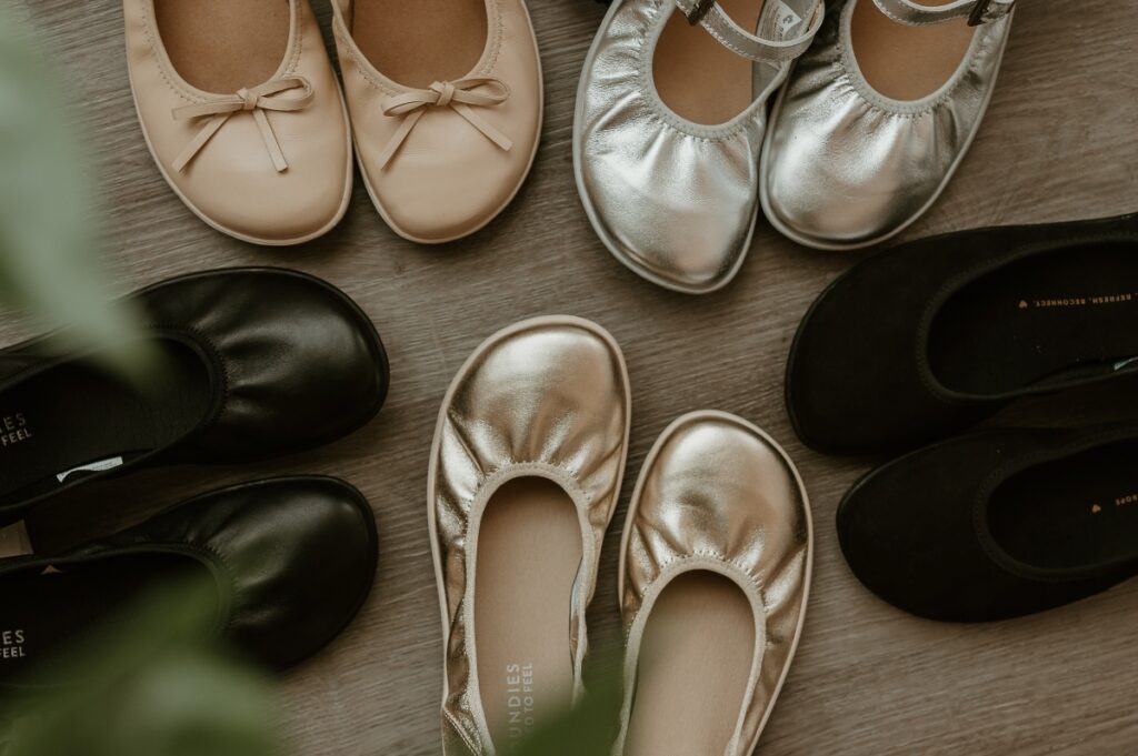 A group of different metallic and neutral ballet flats all sitting with their toes pointing in. The wide toe boxes are displayed in the center of the image.