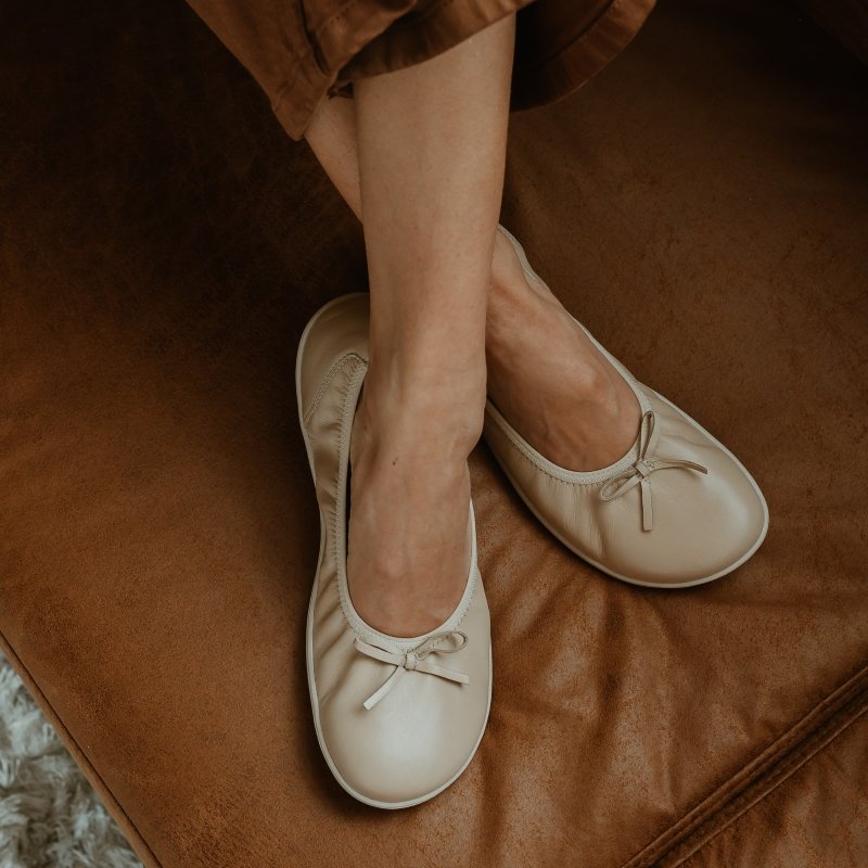 A close up of a persons crossed feet wearing Groundies Ruby Beige leather ballet flats against a cognac colored couch. The wide toe box flats have a dainty bow on the front.
