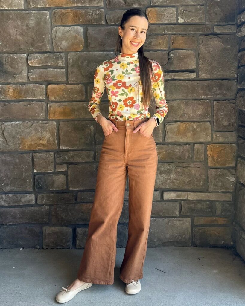 Full body view of a woman in a fun floral top and light brown pants smiling at the camera with her hands in her pockets. She finished the outfit with Groundies Ruby Beige barefoot ballet flats.