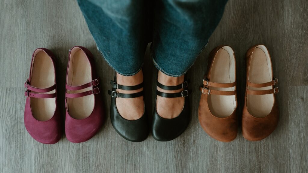 A line of 3 colors of the same Jenon Leather Villet flats with a double strap. There is a person standing in the center pair of black barefoot ballet flats, and the maroon and brown Jenon Villet are on either side.