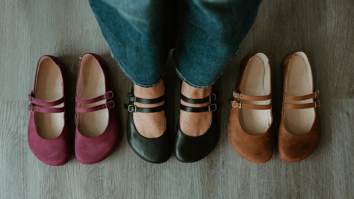 A line of 3 colors of the same Jenon Leather Villet flats with a double strap. There is a person standing in the center pair of black barefoot ballet flats, and the maroon and brown Jenon Villet are on either side.