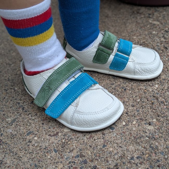 A pair of white leather sneakers from Magical Shoes with colorful velcro straps being worn by a toddler in mismatched striped socks