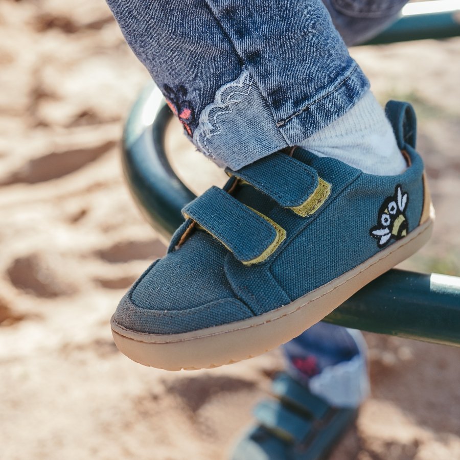 Close up of a pair of blue green canvas barefoot Mukishoes sneakers with an embroidered bee near the ankle. The lightweight flexible outsole is being used on playground equipment