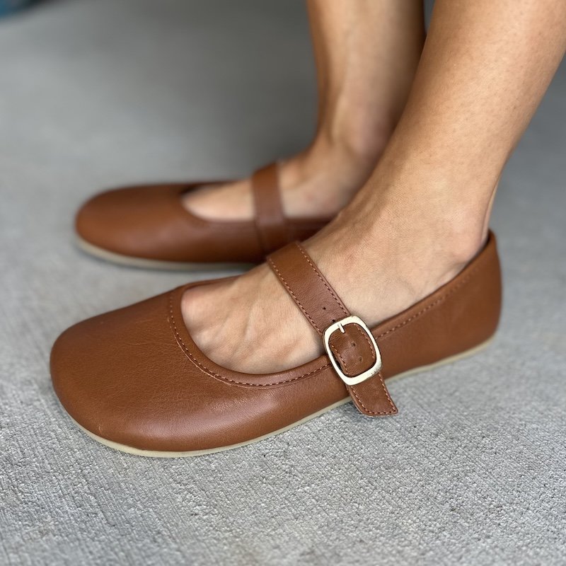 A close up side view of a pair of feet standing on concrete wearing Origo Shoes barefoot mary janes with a single strap in cognac brown leather