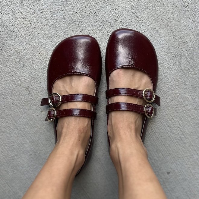 A top down view of a pair of feet standing on concrete wearing Origo Shoes burgundy two strap wide toe box mary janes
