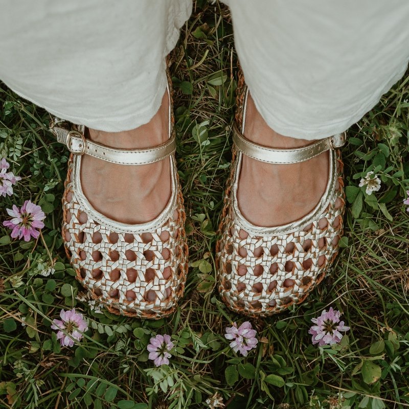 Top down look at a pair of Gold woven leather ballerinas from Origo Shoes. Beige pants are visible hovering over the shoe and purple flowers are on the ground. The wide anatomical toe box is prominently shown in the middle of the photo