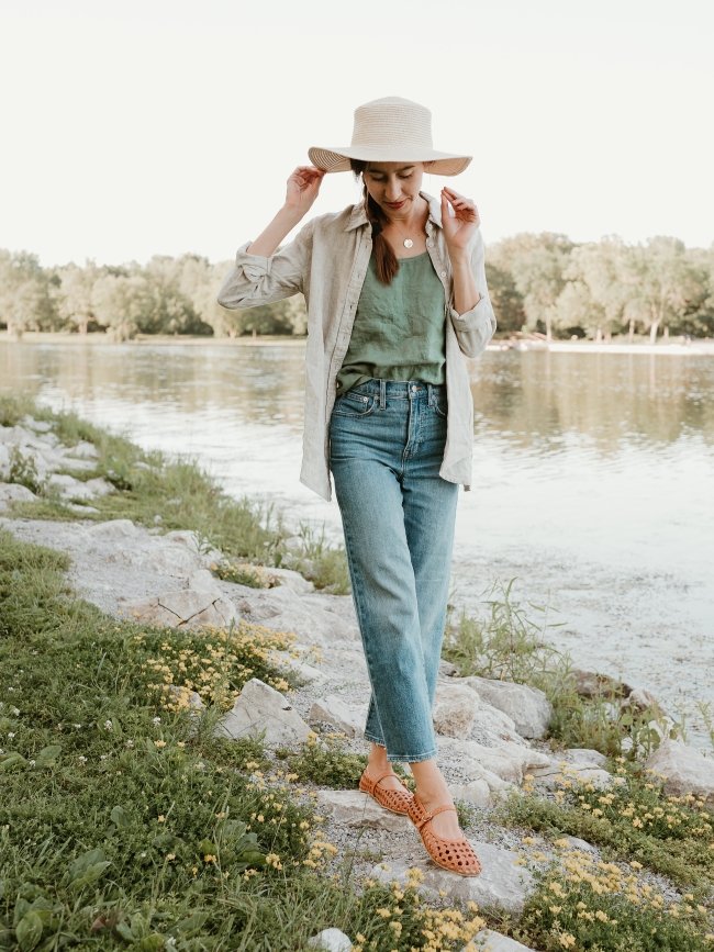 Full body photo of a woman in a sun hat, jeans, and breeze linen layers looking down at her feet while she walks along a rocky beach covered in tiny yellow flowers. She has cognac woven leather ballerinas from Origo Shoes with a wide toe box and zero drop flexible sole on her feet.