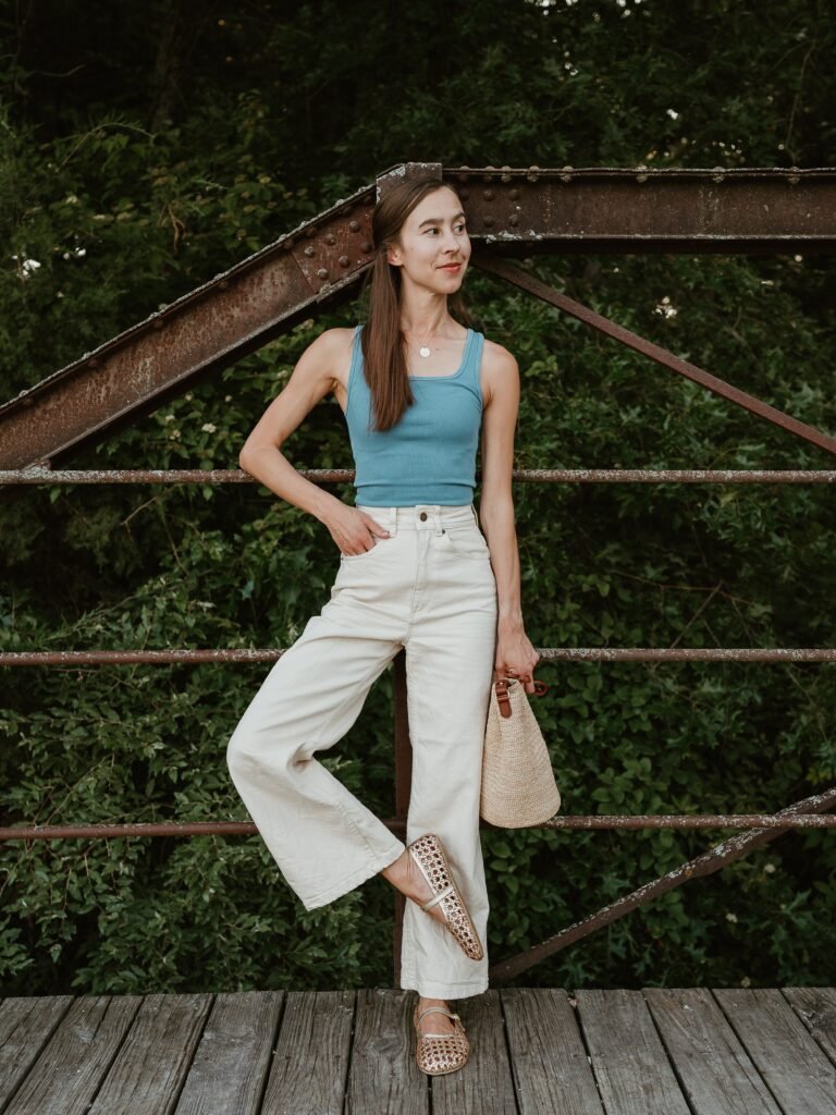 Full body image of a woman with long brown hair leaning against a rusty bridge. She is wearing wide leg beige pants and a robins egg blue tank. She has one foot crossed in front of the other and her feet are adorned with Origo Shoes Woven Ballerinas in gold leather with a wide comfortable toe box