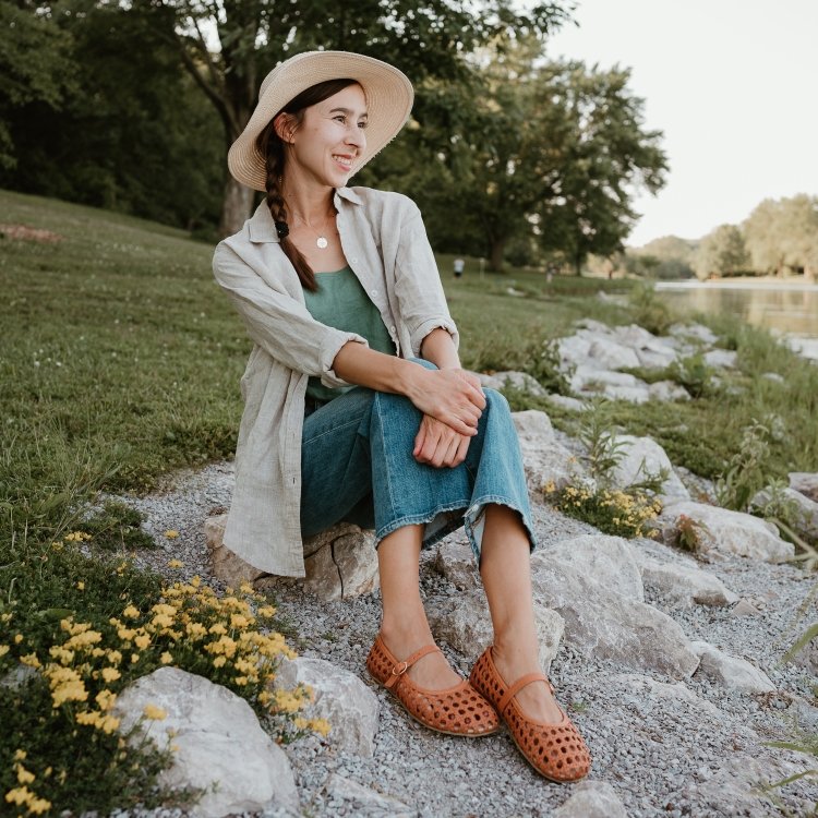 A woman in summery breezy casual clothes sitting on rocks on the shore of a lake, staring off into the distance with a smile on her face. She has Origo Woven leather ballerinas barefoot flats on her feet