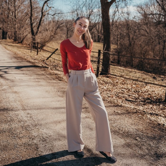 Full body view of a woman standing outside on a dirt path. She is wearing a burnt orange long sleeved shirt, beige wide leg dress pants, and Peerko Petite barefoot flats in Bonnie Plaid.