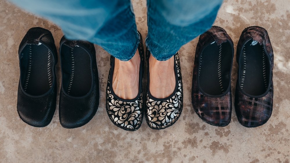 A line up of Peerko Petite barefoot ballet flats in 3 different colors. There is a person wearing the middle swirly silver pair while a textured black pair and Bonnie Plaid pair sit on the sides.